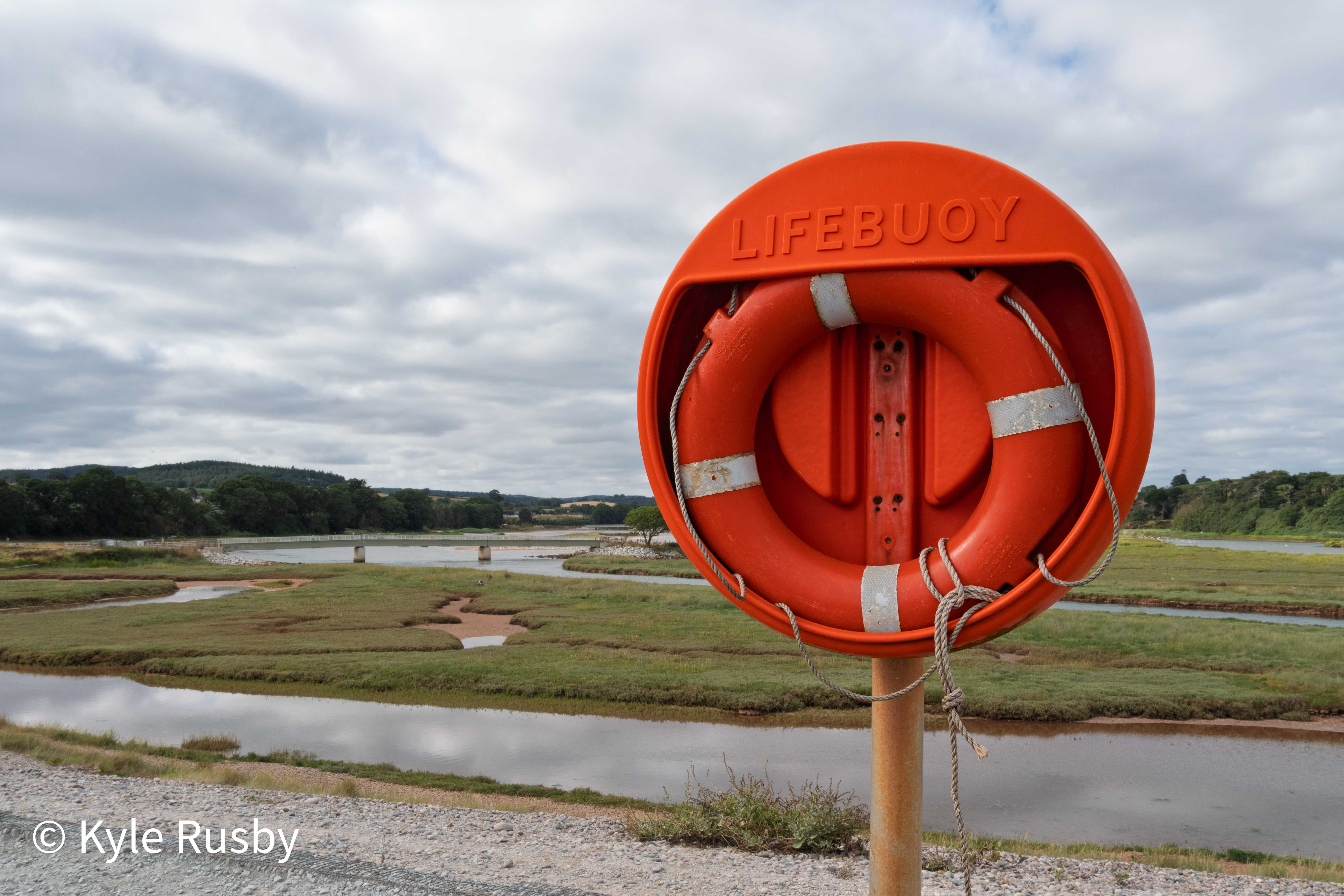 Bright orange lifebuoy mounted on a post overlooking the salt marshes and winding channels of the River Otter estuary under a heavy grey sky