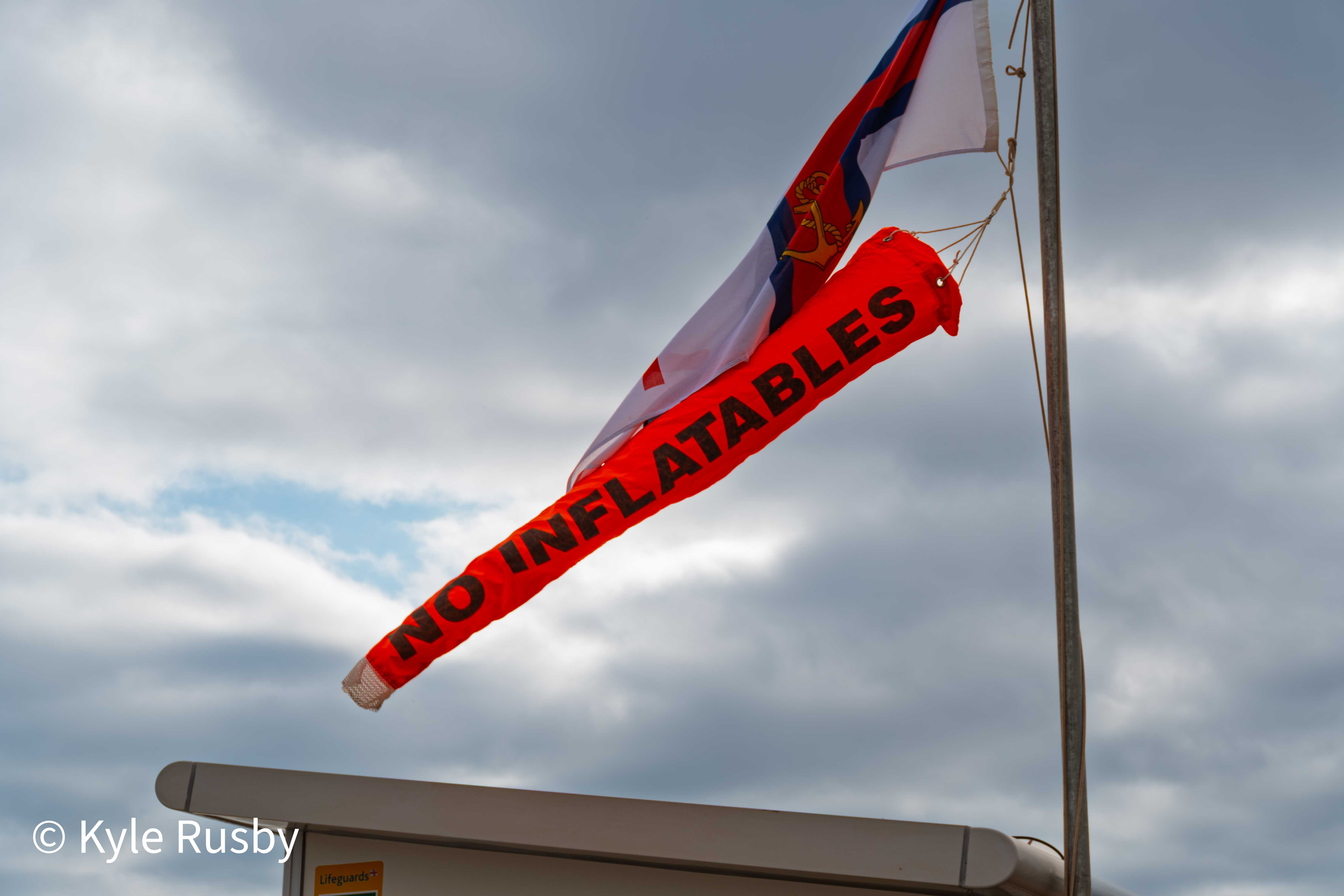A red No Inflatables windsock and a Royal Navy ensign flying from a seafront flagpole against dark clouds