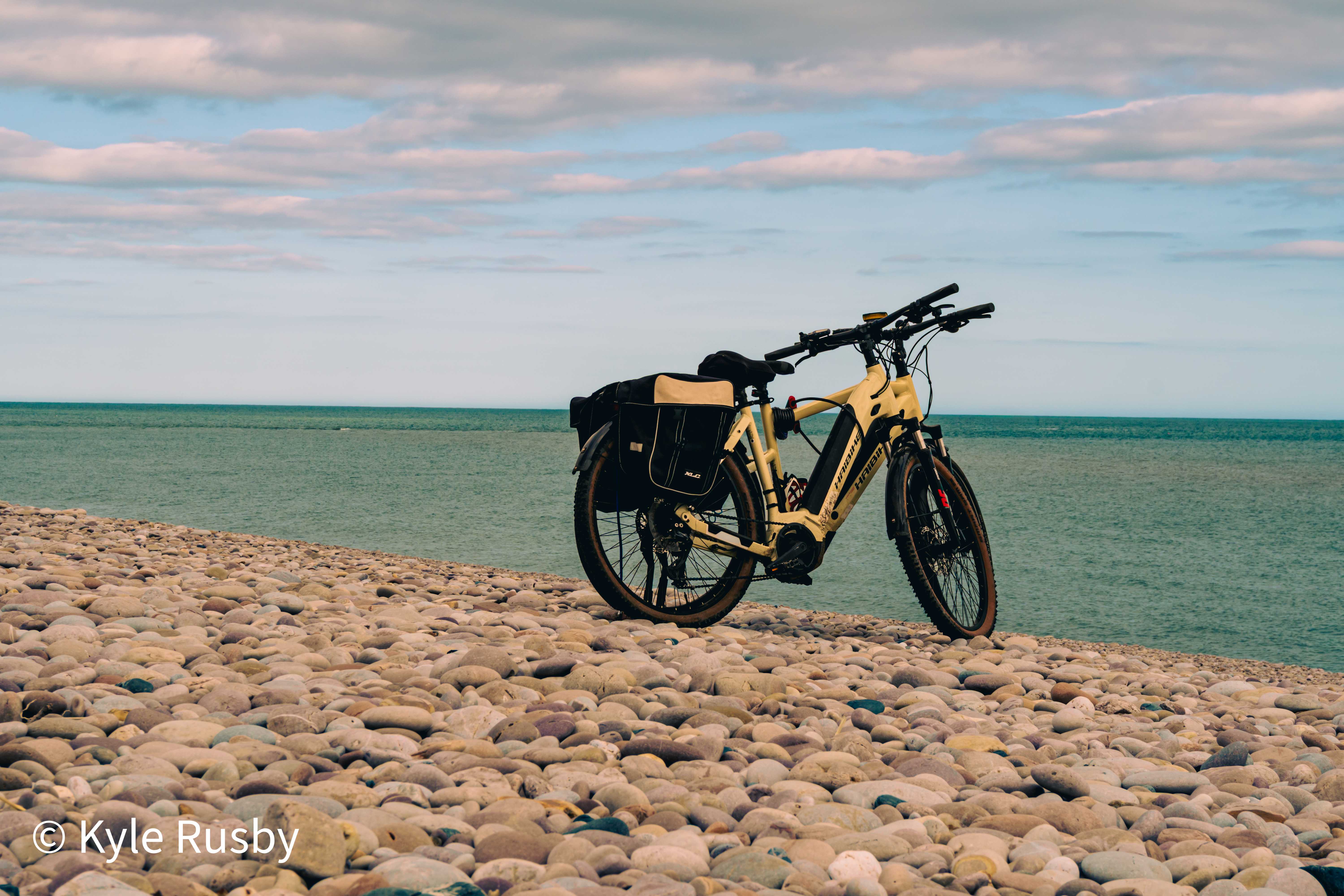 Two touring e-bikes with panniers propped together on a steep bank of pink and grey pebbles, with a calm teal sea behind