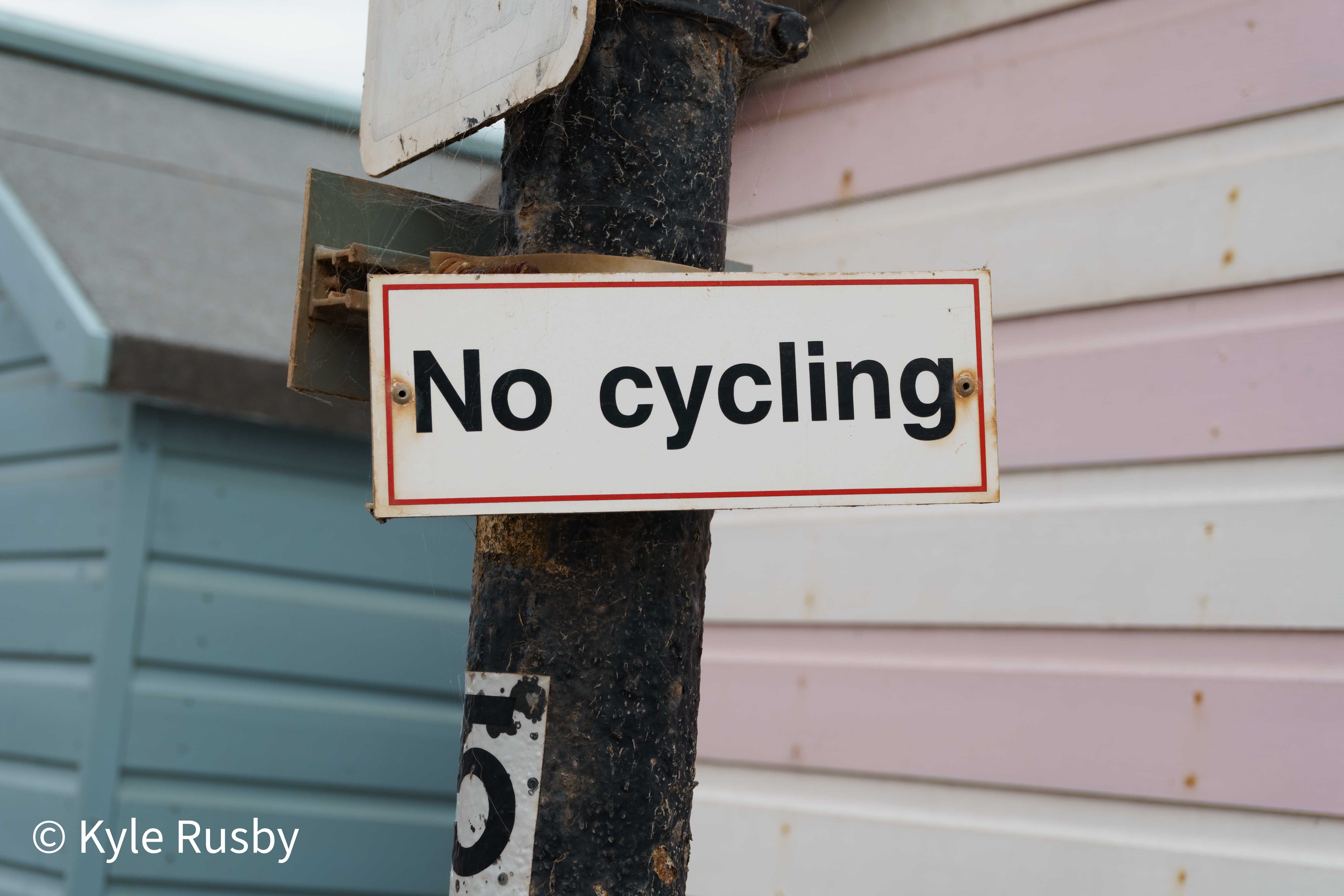 A white enamel No Cycling sign bolted to a weathered black post in front of pastel pink and blue painted beach huts