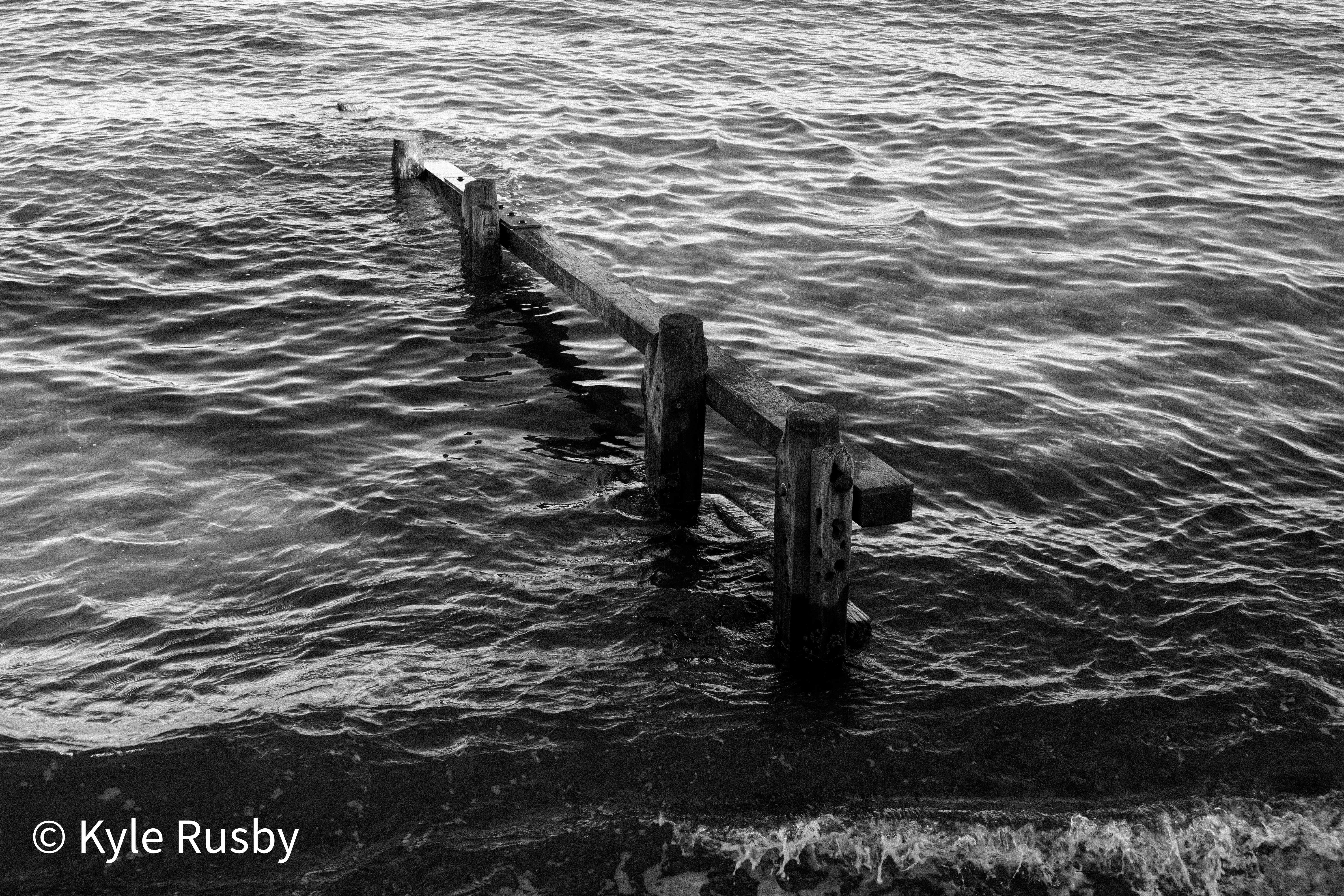 Black and white photograph of an old wooden groyne running out into dark rippled water, waves breaking at its landward end