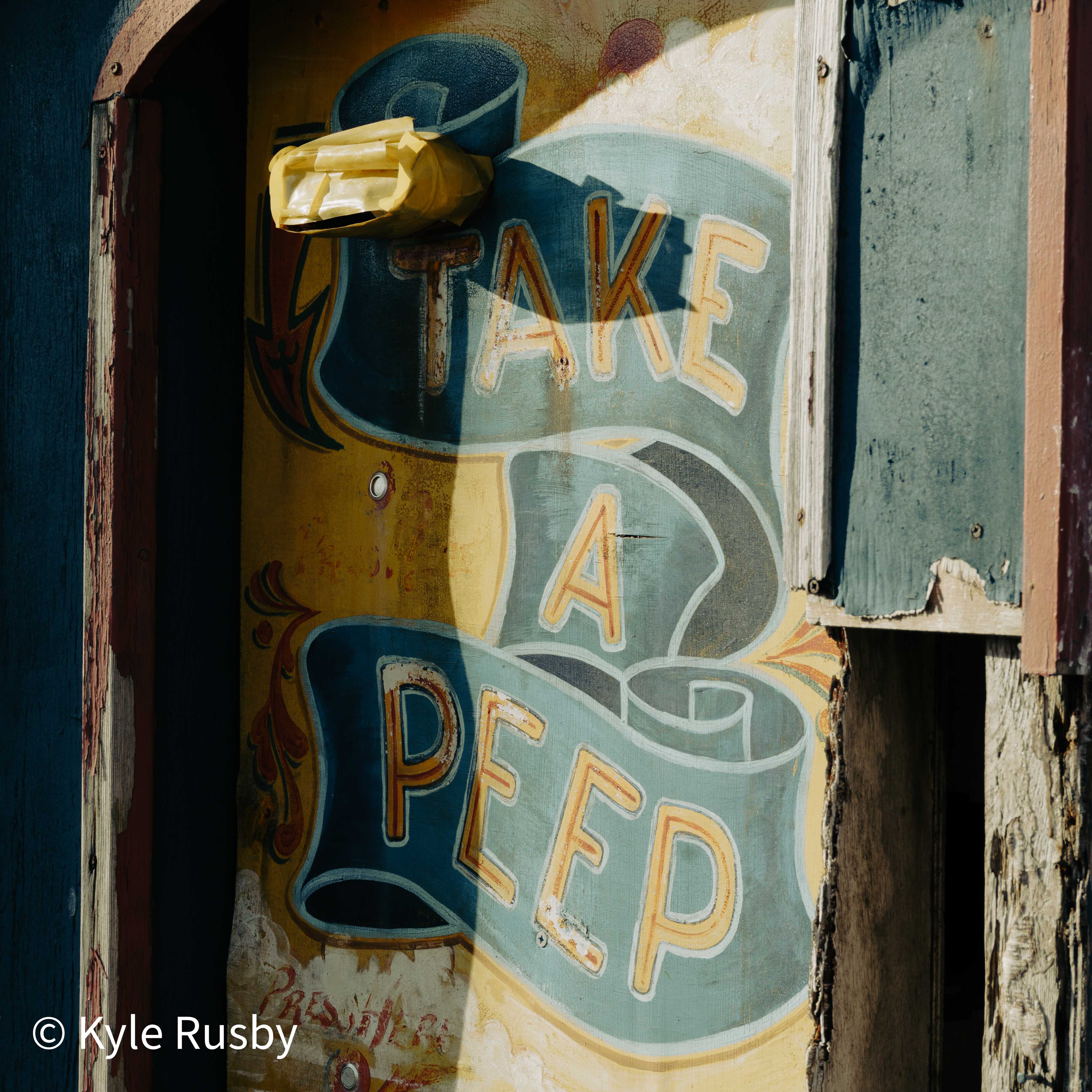 A weathered fairground-style sign painted with "Take a Peep" on blue ribbon scrolls, set in a peeling wooden hut door in hard sunlight