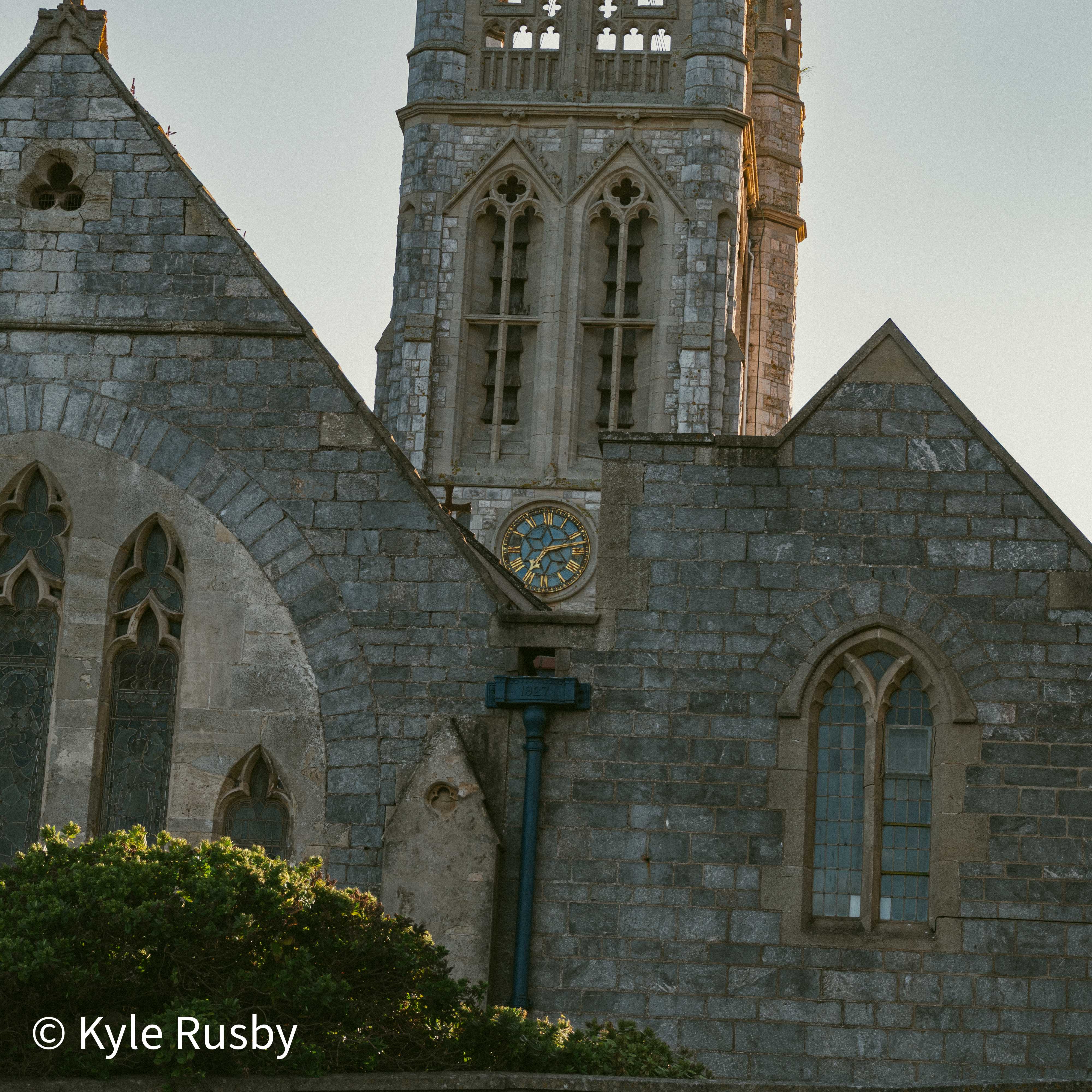 A grey stone Victorian church in low evening light, its tower and clock rising between two pitched gables with arched windows