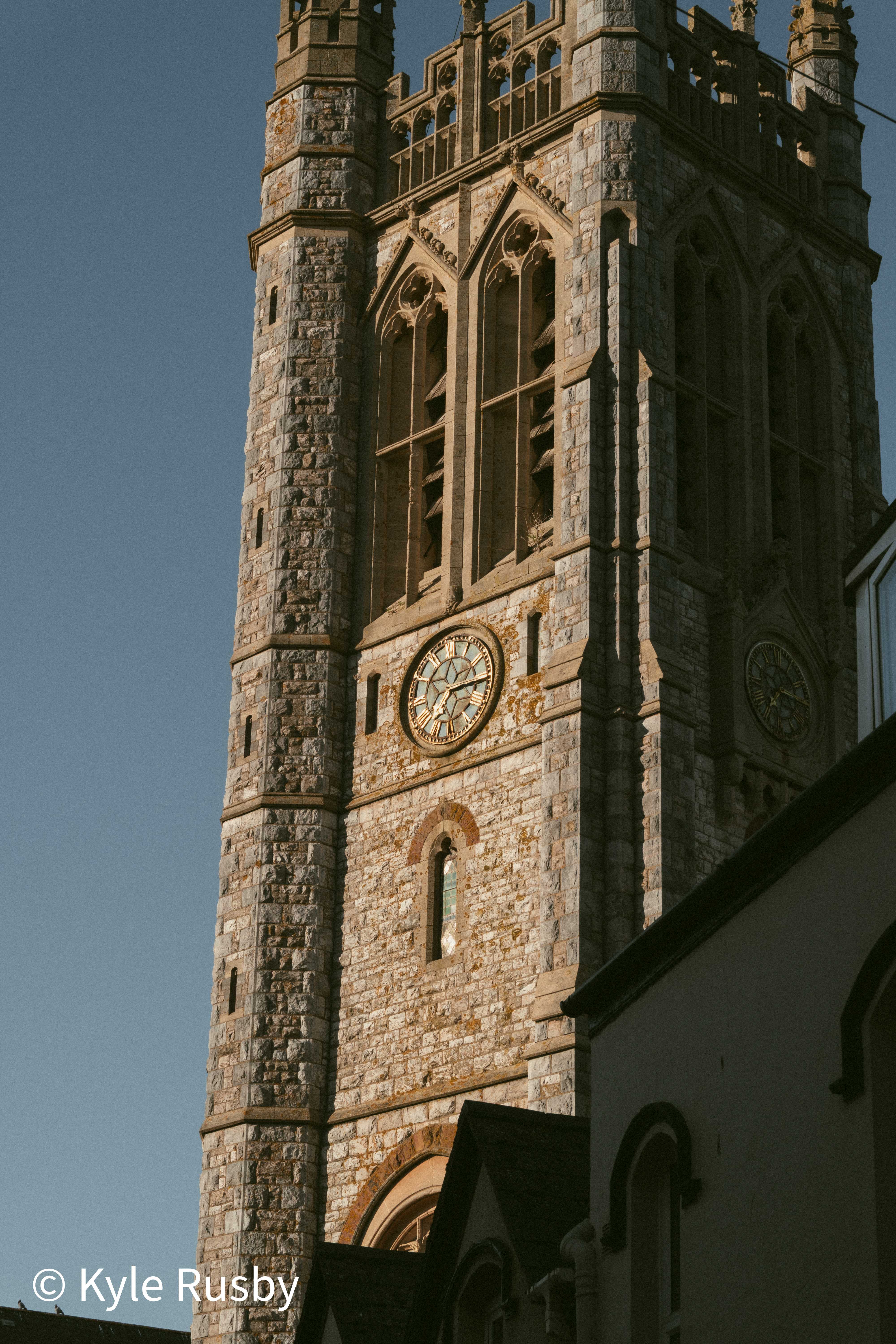 A tall gothic church tower with pinnacles and a pale clock face, half in shadow against a clear evening sky