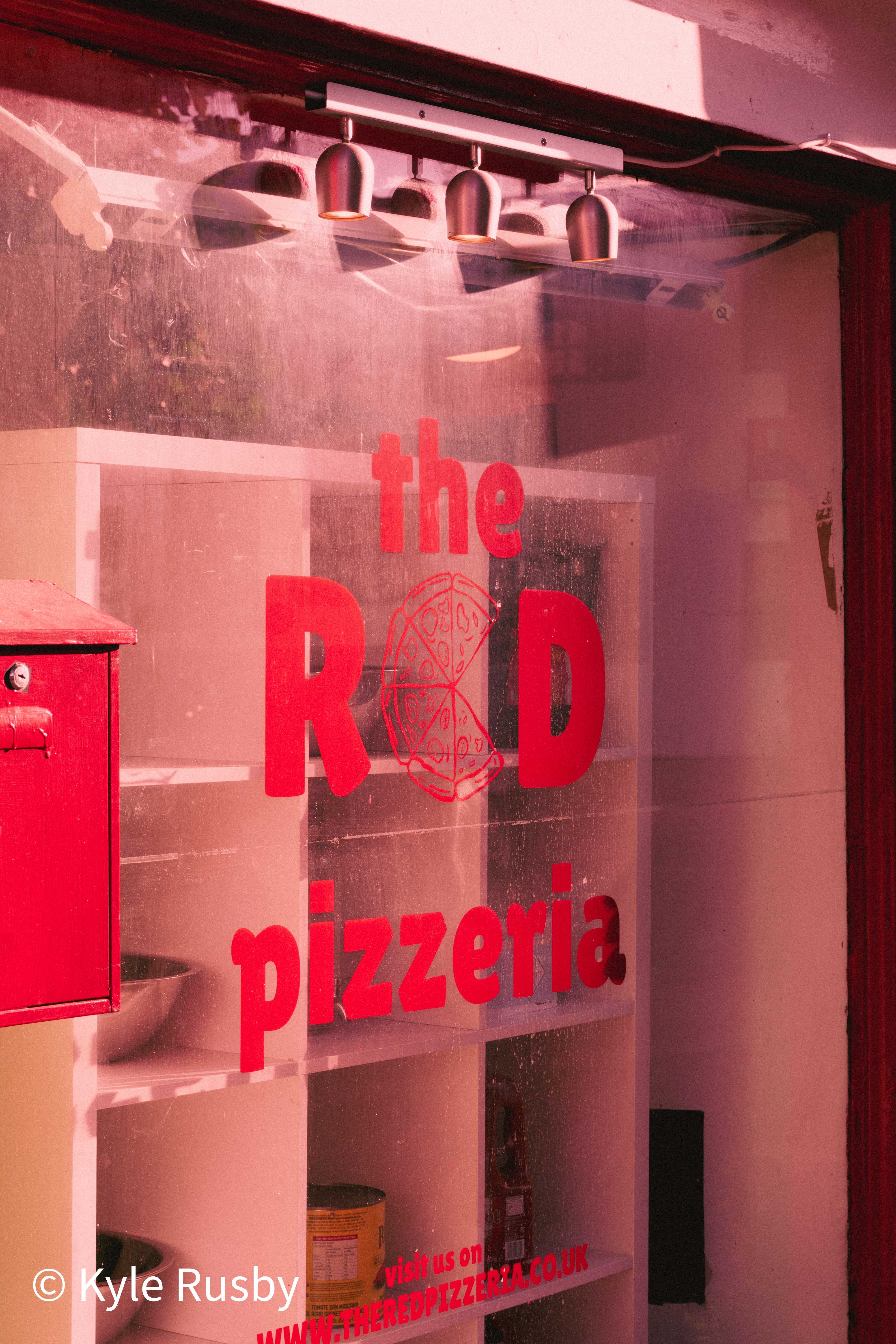 A shop window lettered "the RED pizzeria" in bright red, the whole scene washed in pink light, empty white shelves and a red postbox inside