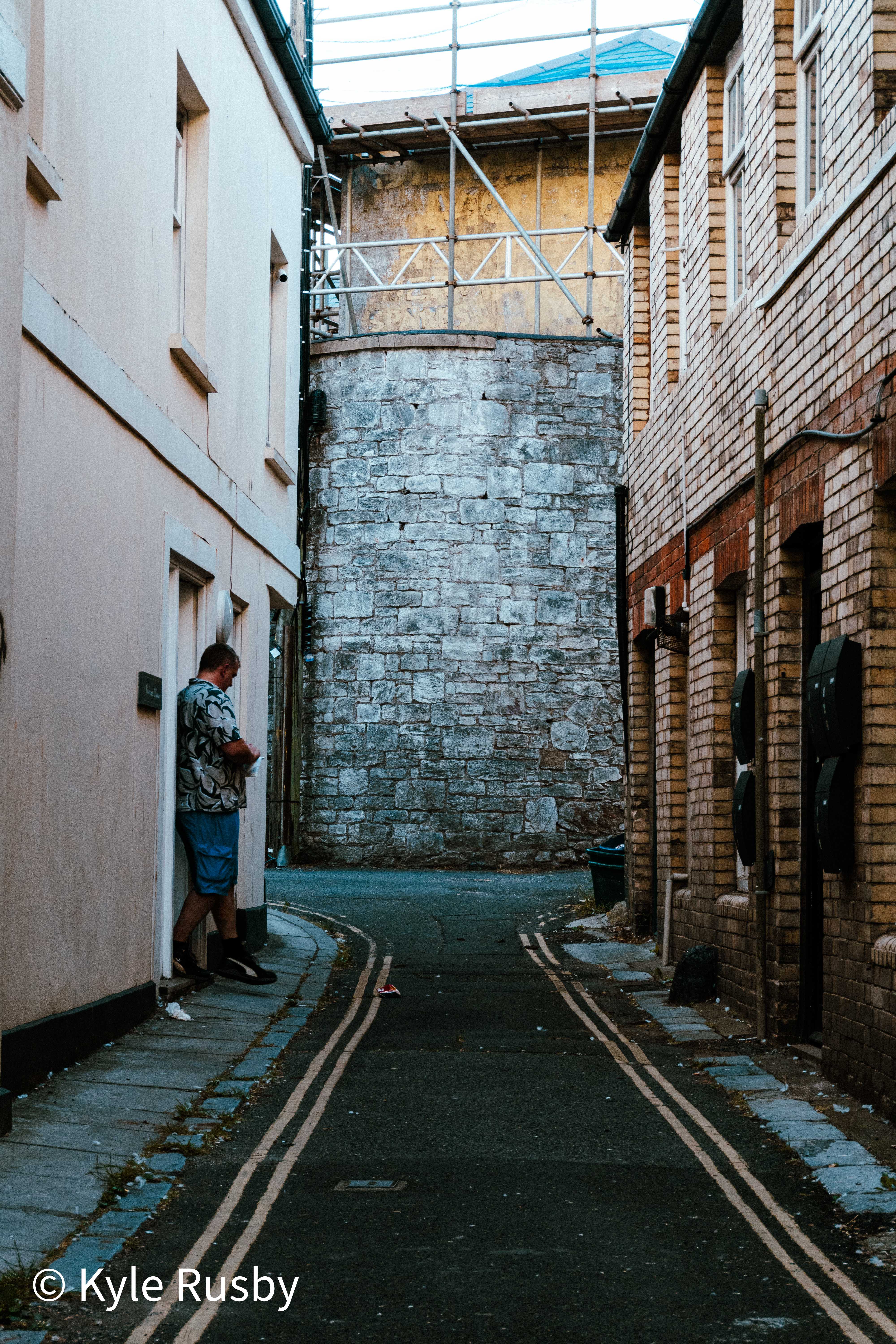 A man leans against a wall checking his phone in a narrow back lane of painted and brick houses, a round stone tower under scaffolding at the end