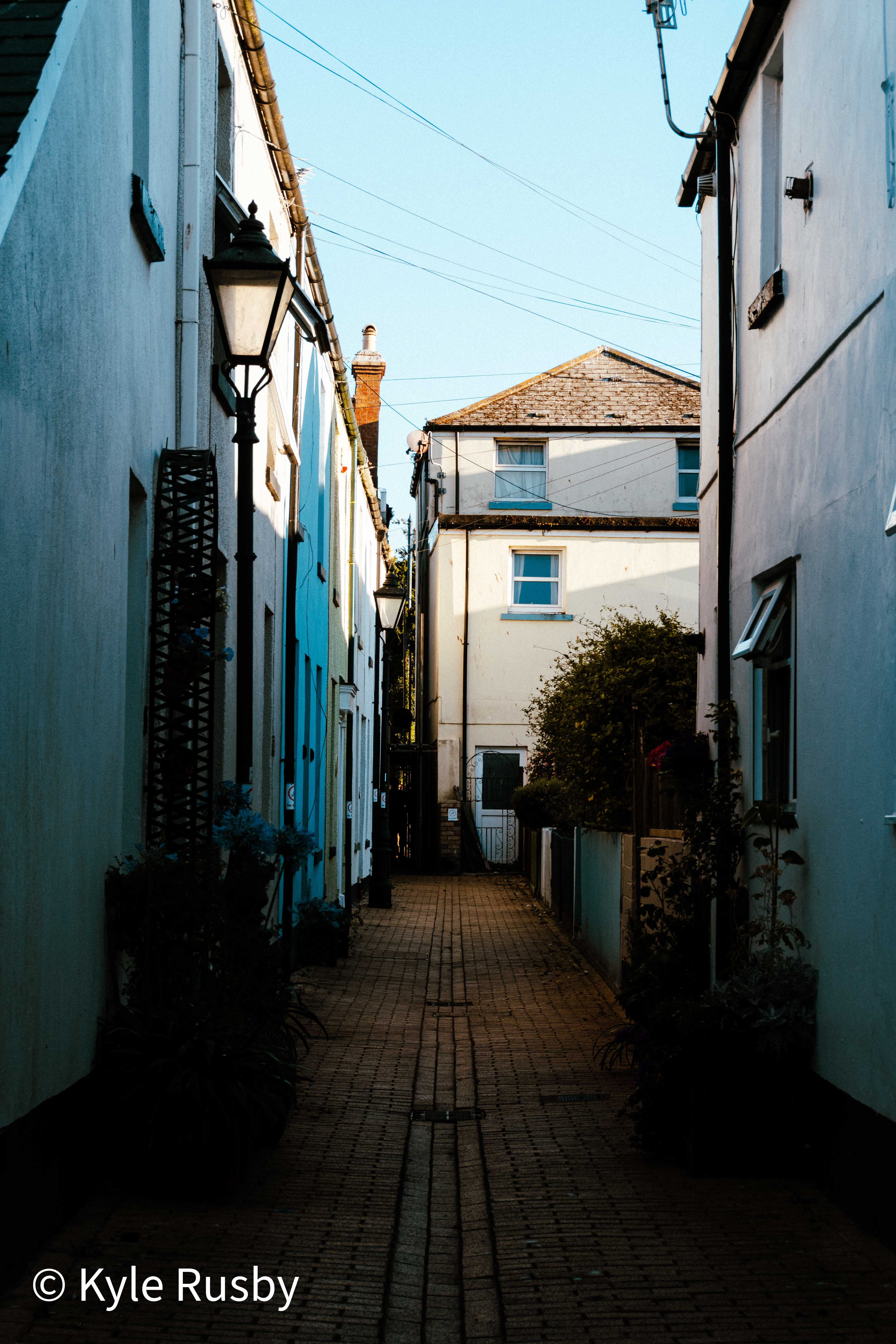 A narrow brick-paved passage between pastel cottages in low sun, a lamp post halfway down and washing lines crossing the sky