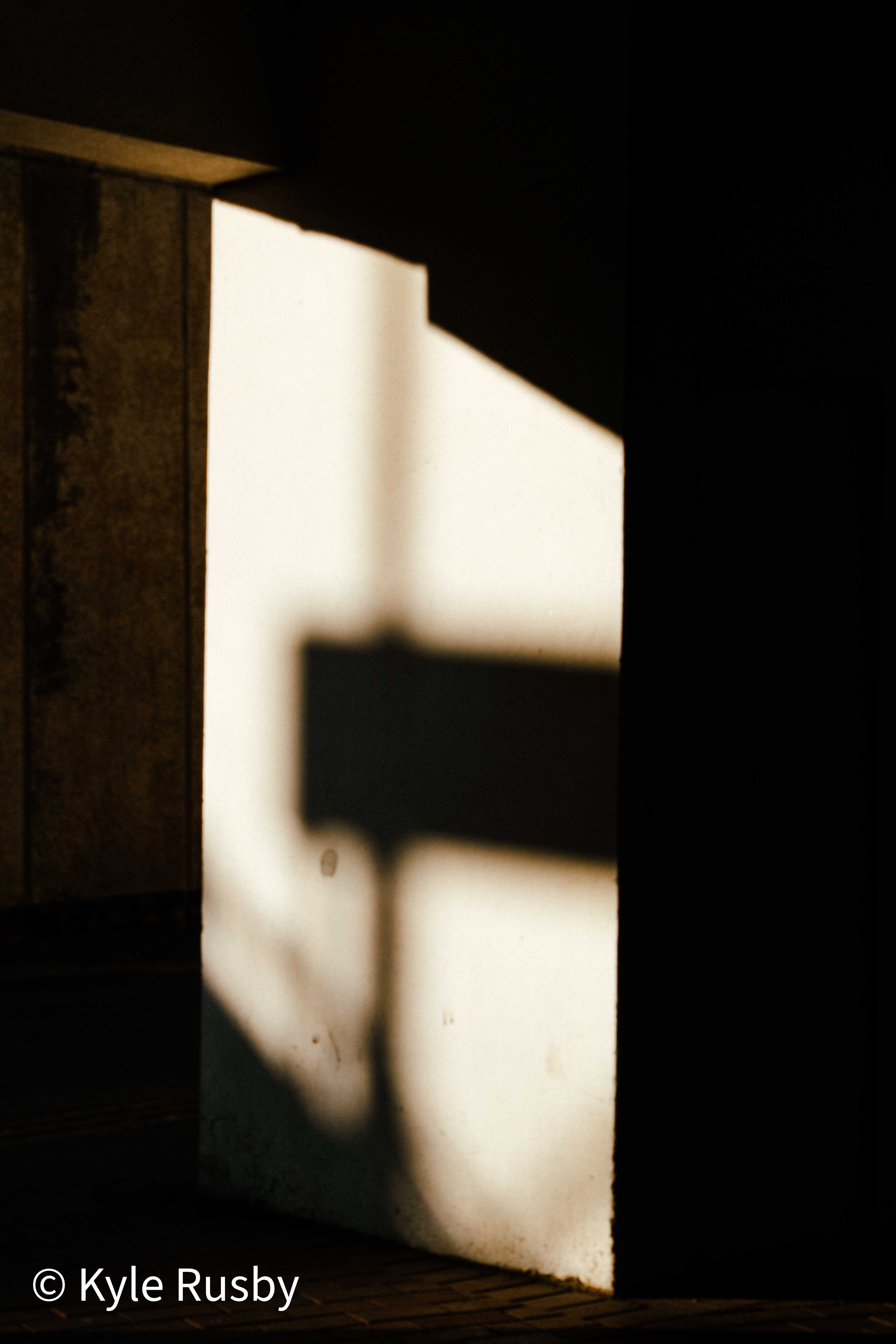 Hard evening light throws the shadow of a sign bracket across a pale wall framed by deep black shadow