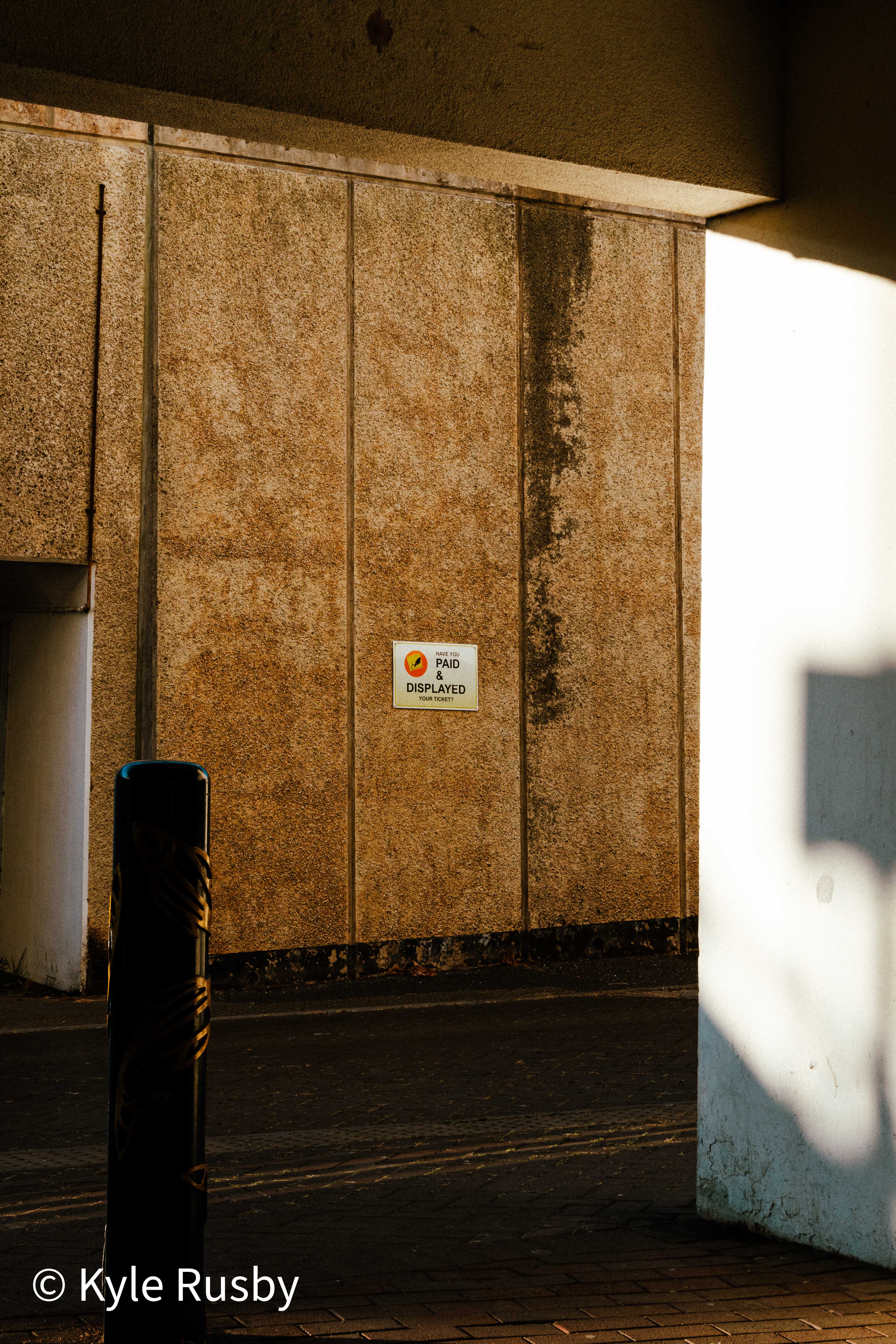 A small Pay & Display sign on a stained pebbledash wall in raking evening light, half the frame falling into shadow