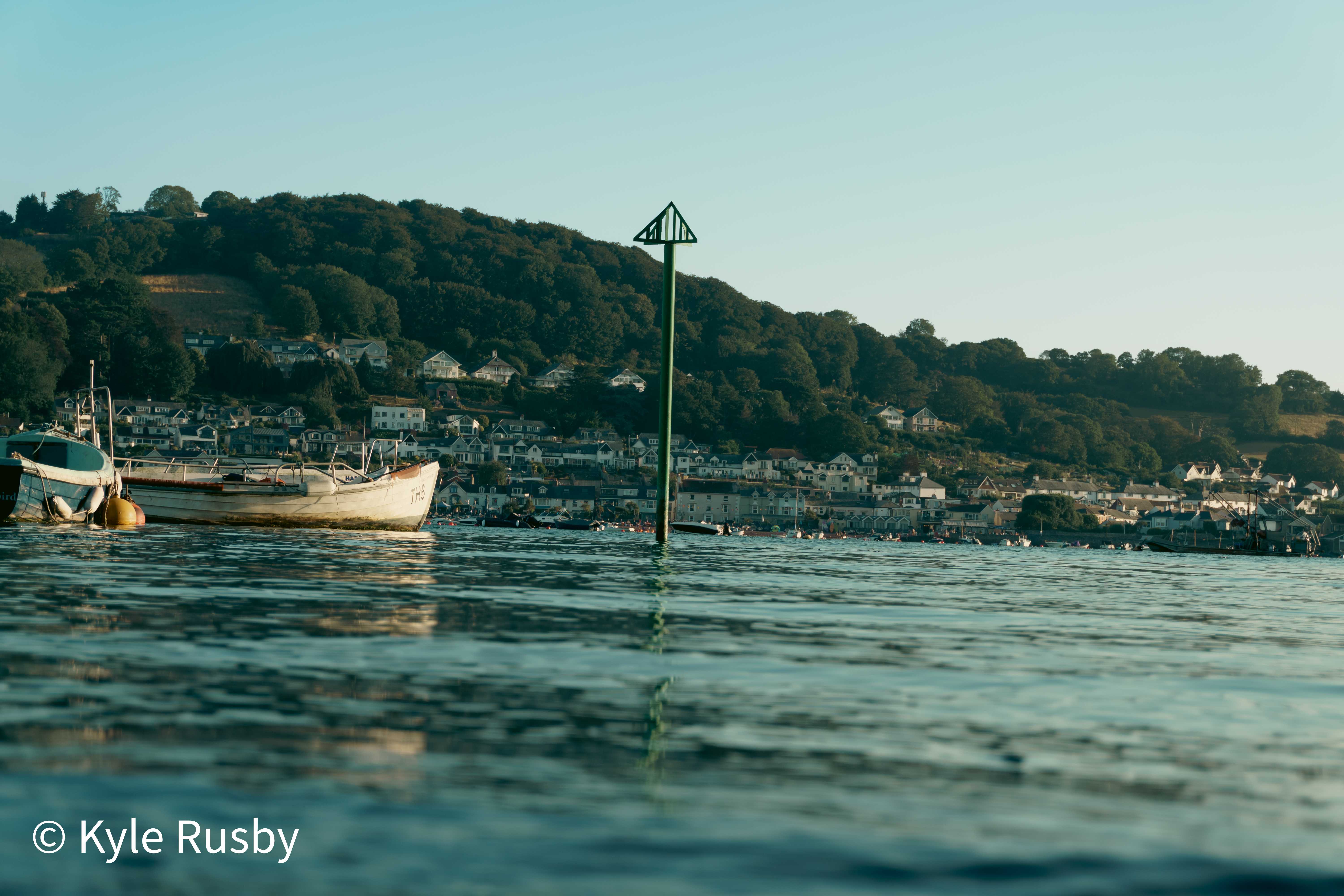 Seen from water level in golden evening light: a green tide marker and a small moored boat on calm water, a hillside town of white houses rising behind