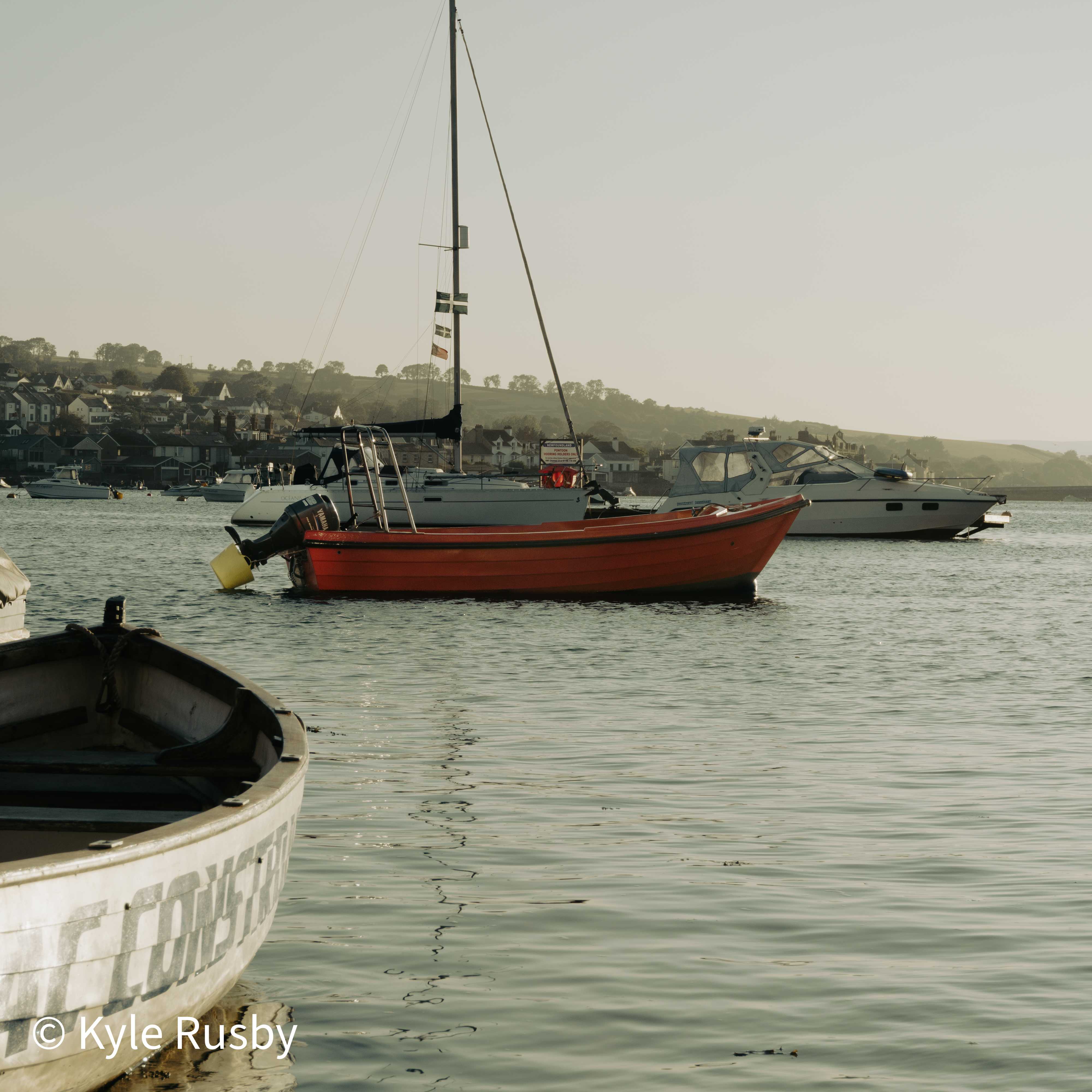 An orange wooden boat moored among sailboats and cruisers on a hazy evening estuary, a Devon flag on the mast and the town rising on the far shore