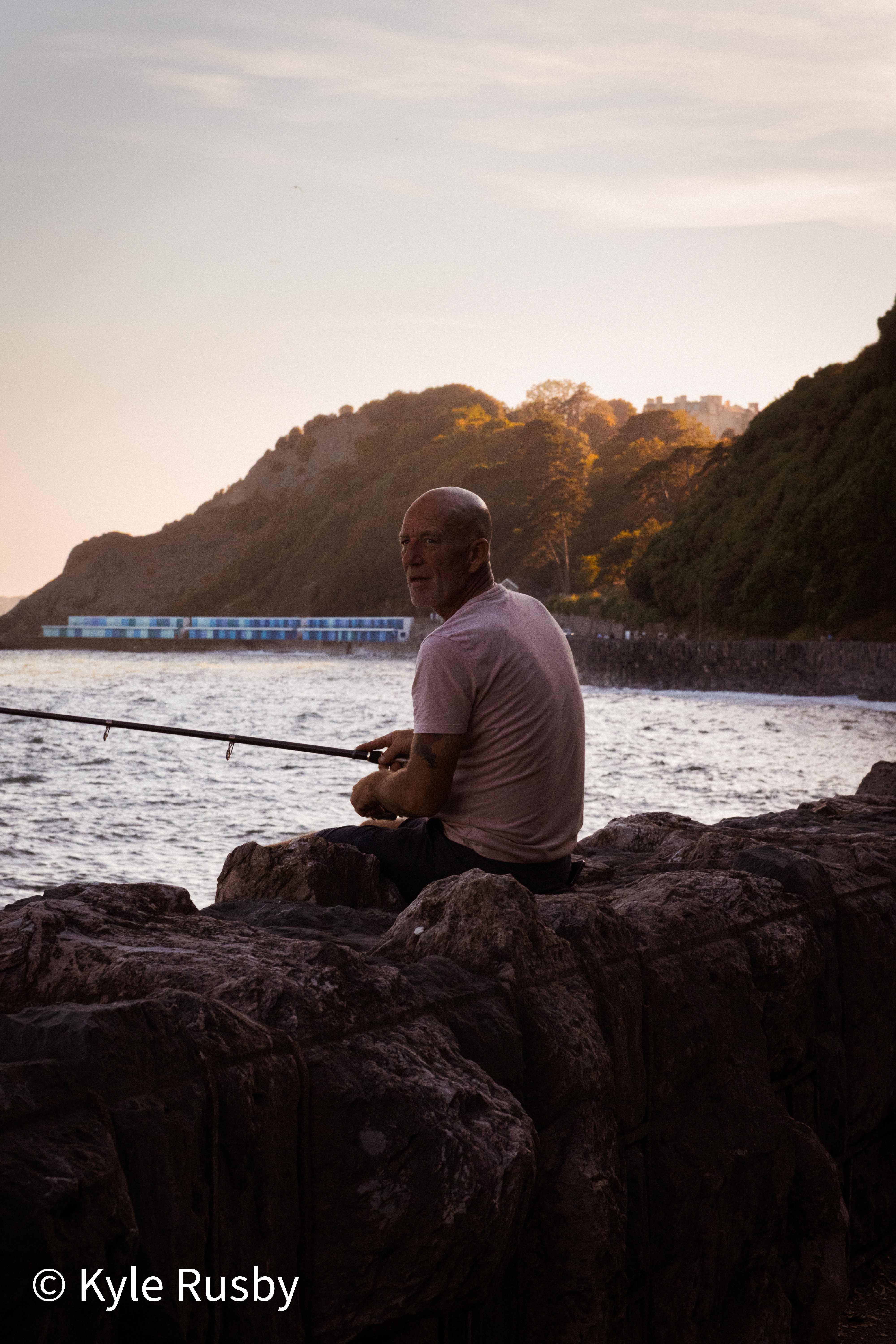 The fisherman turns from his rod to glance back over his shoulder, the headland and huts holding the last light behind him