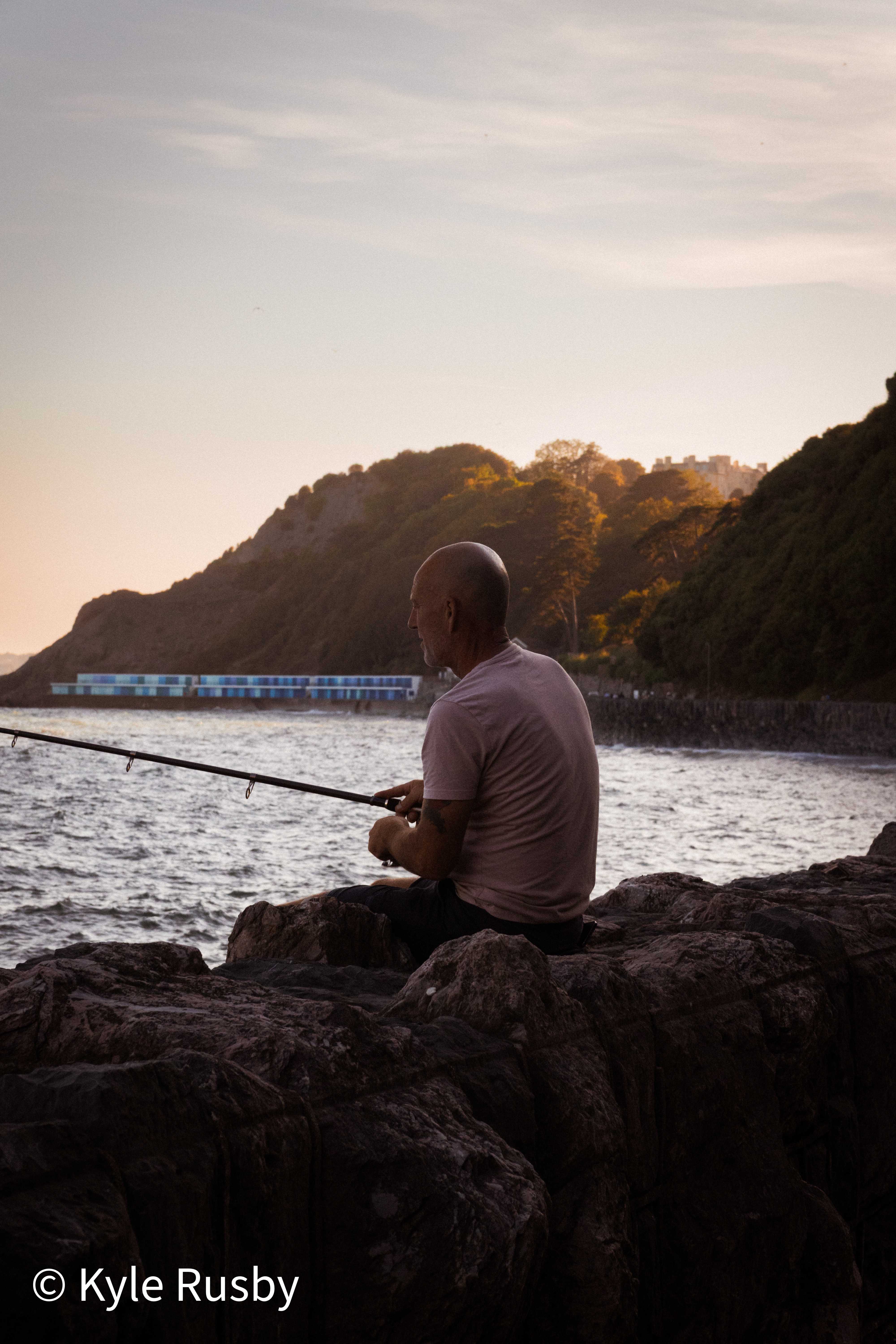 A fisherman sits on the rocks at dusk with his rod out over the water, a line of blue beach huts and a wooded headland catching the last sun behind