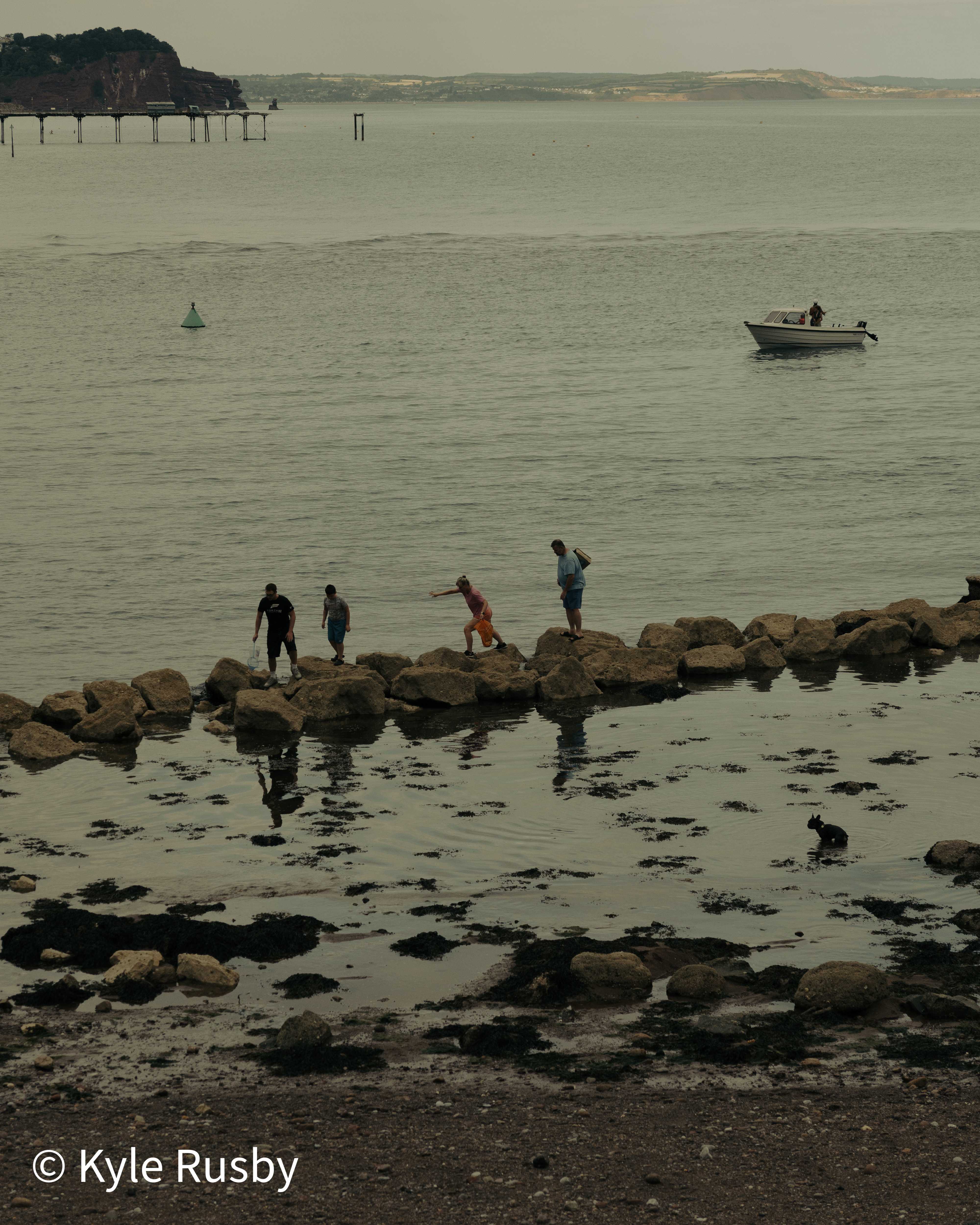 Four people pick their way along a rock breakwater at low tide, an angling boat offshore and a pier and red sandstone headland beyond under a flat grey sky