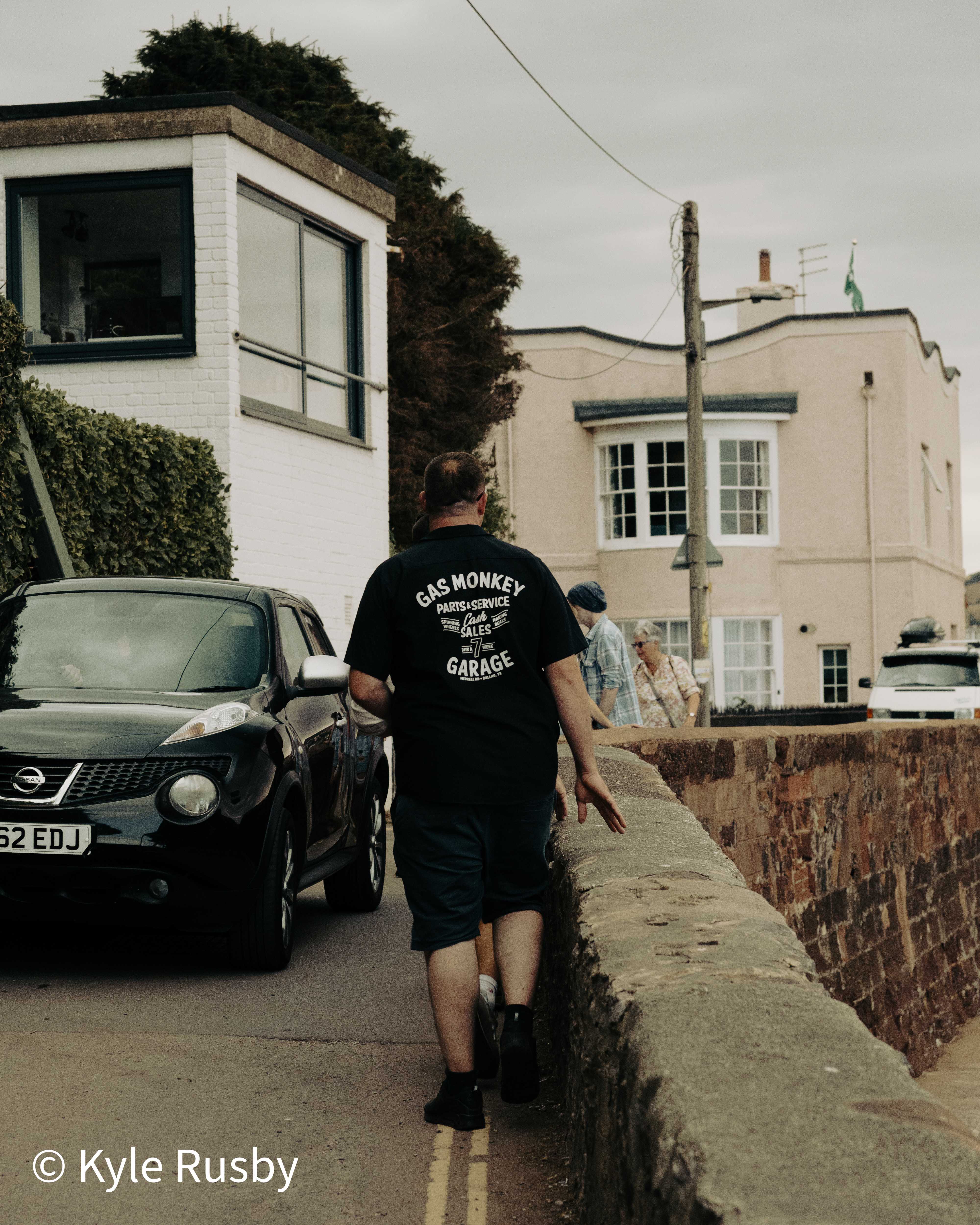 A man in a Gas Monkey Garage t-shirt walks up a narrow seaside lane past a parked black car, a couple coming the other way beside a red sandstone wall