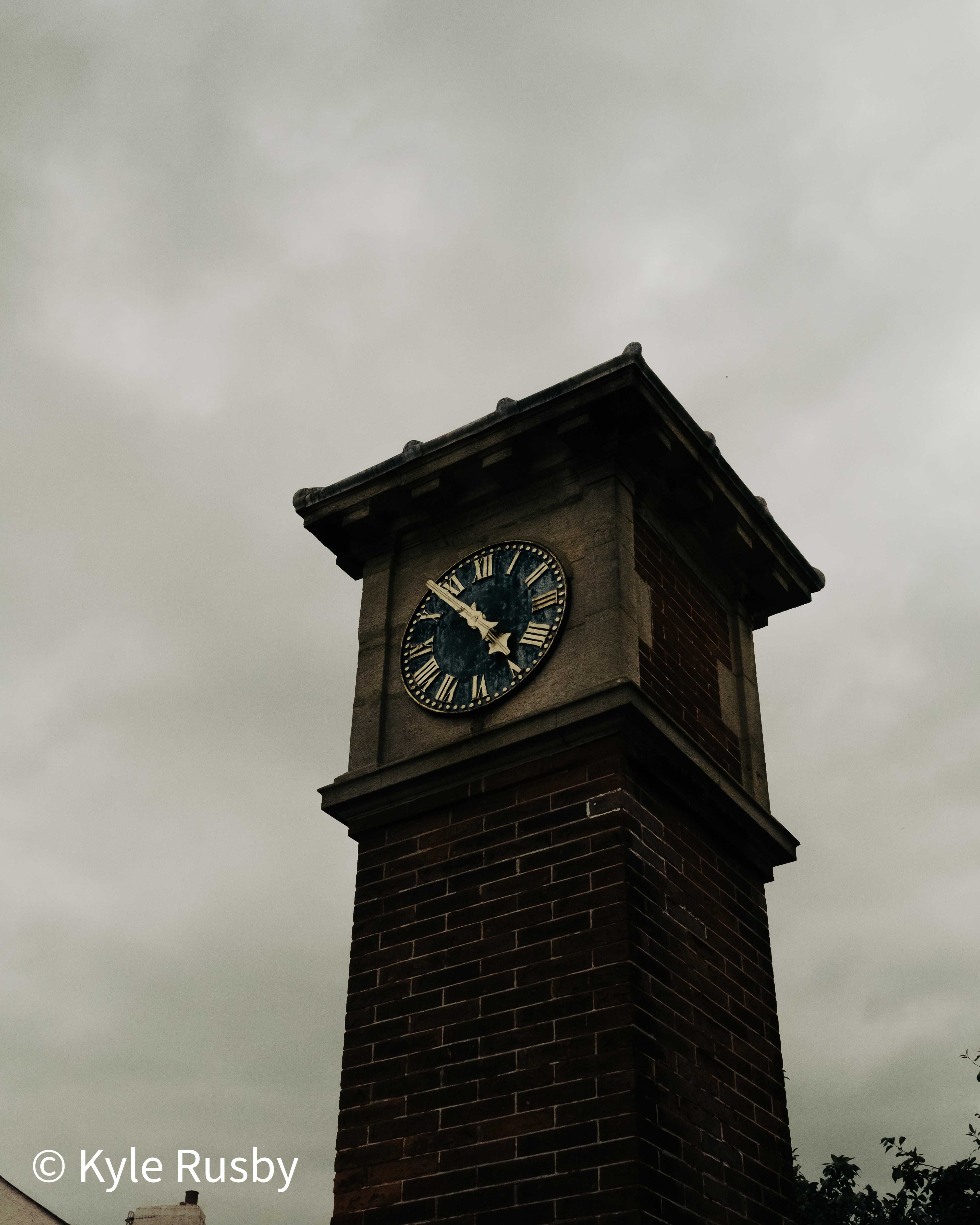 A brick and stone clock tower with a dark Roman-numeral face and gilt hands stands against a flat grey sky