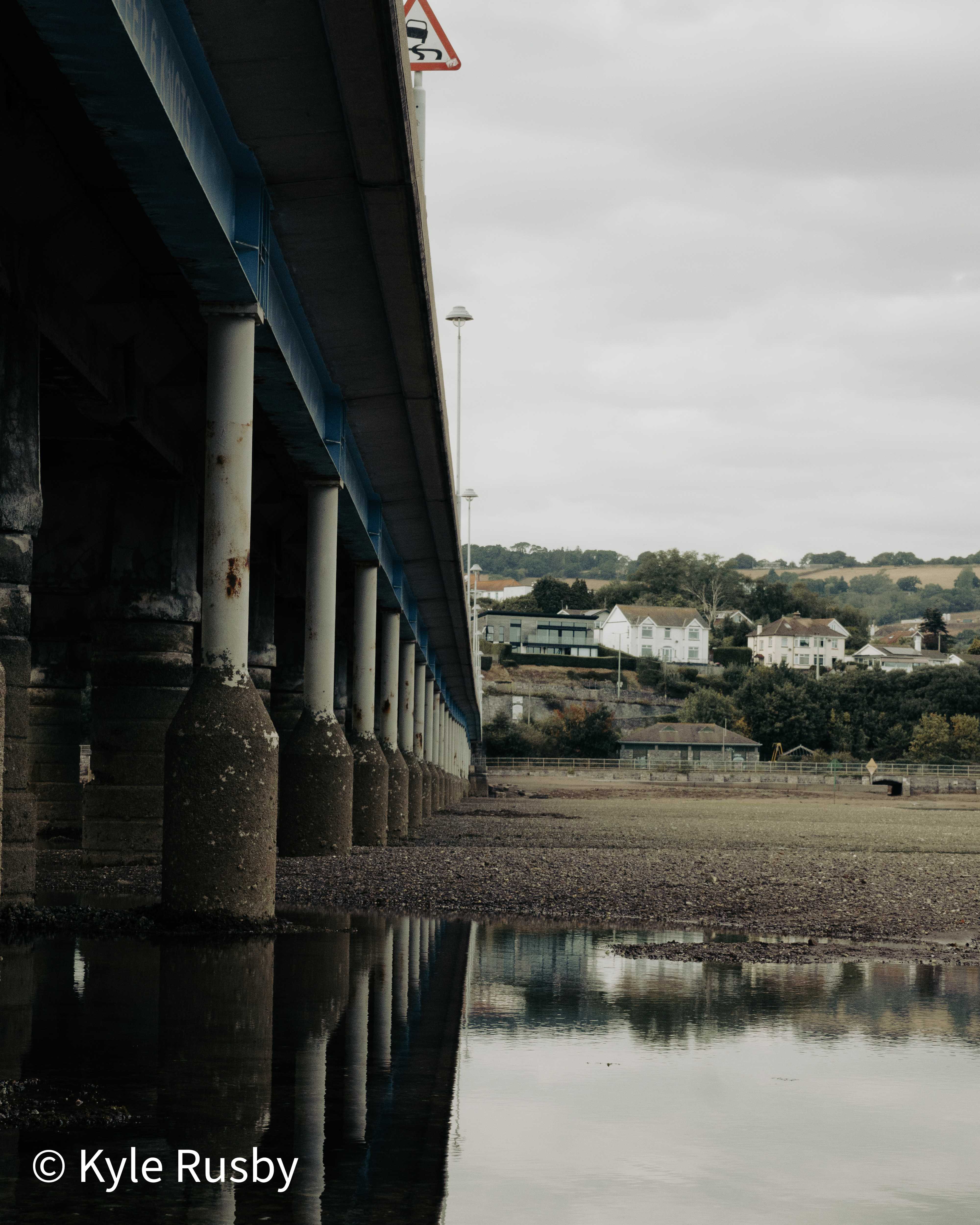 The blue-painted underside of a long road bridge runs away to a vanishing point on its line of concrete piers, low-tide mud and reflections below