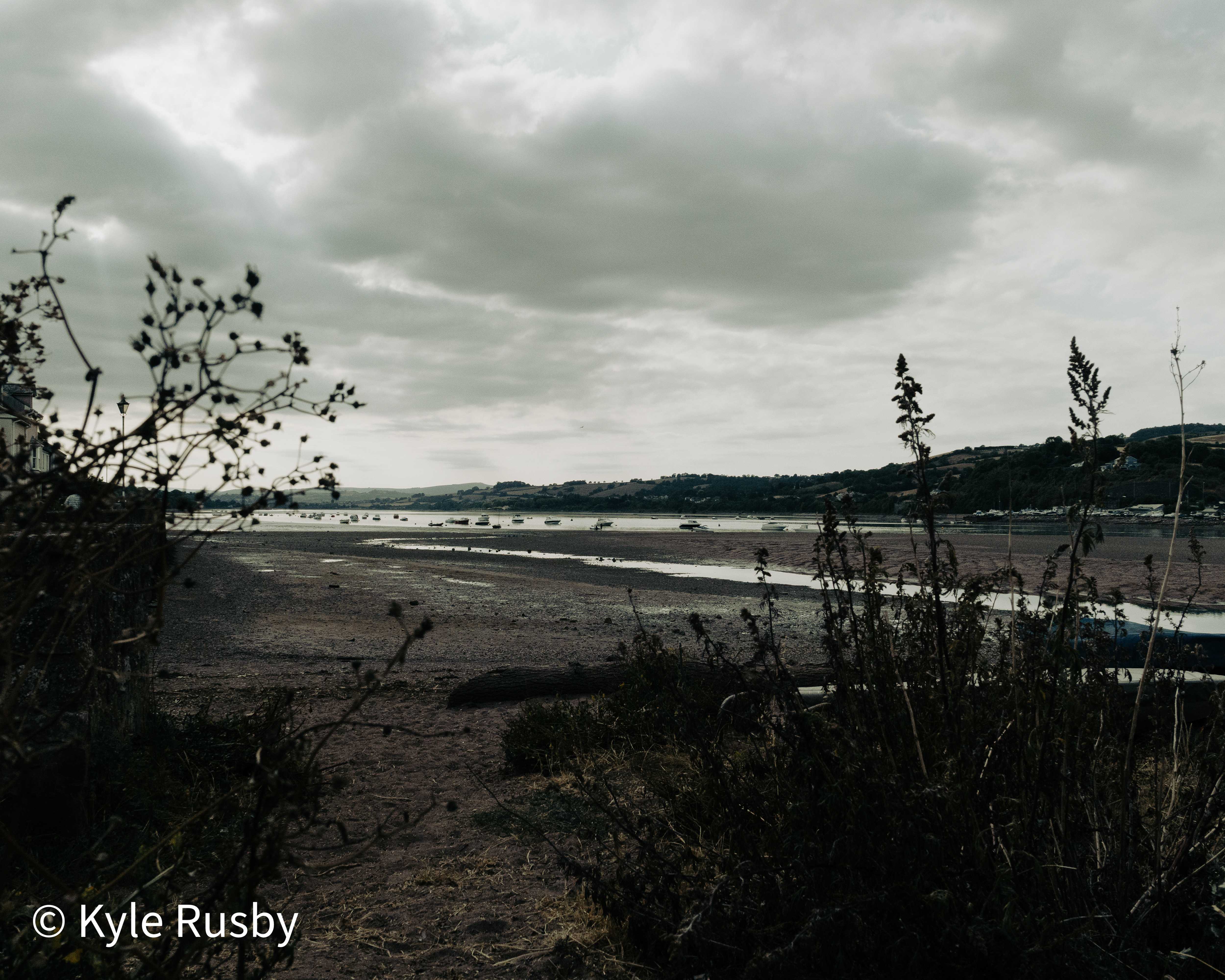 Dead thistles and dry scrub stand along the near shore of an estuary emptied to mud and thin channels, moored boats aground and a heavy cloudbank overhead