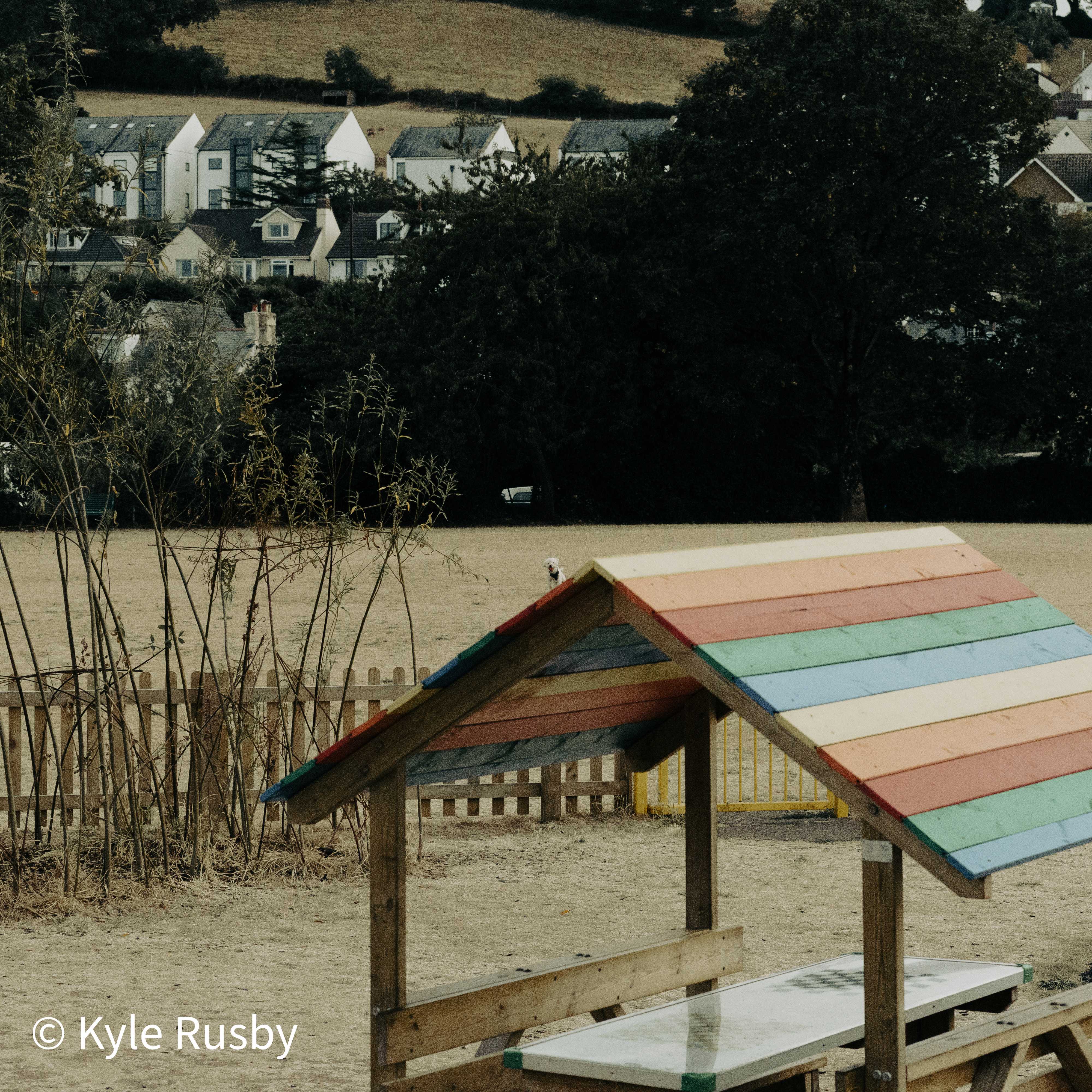 A wooden picnic shelter roofed in painted rainbow stripes stands on parched grass, dry reeds to one side and white houses on the hill beyond