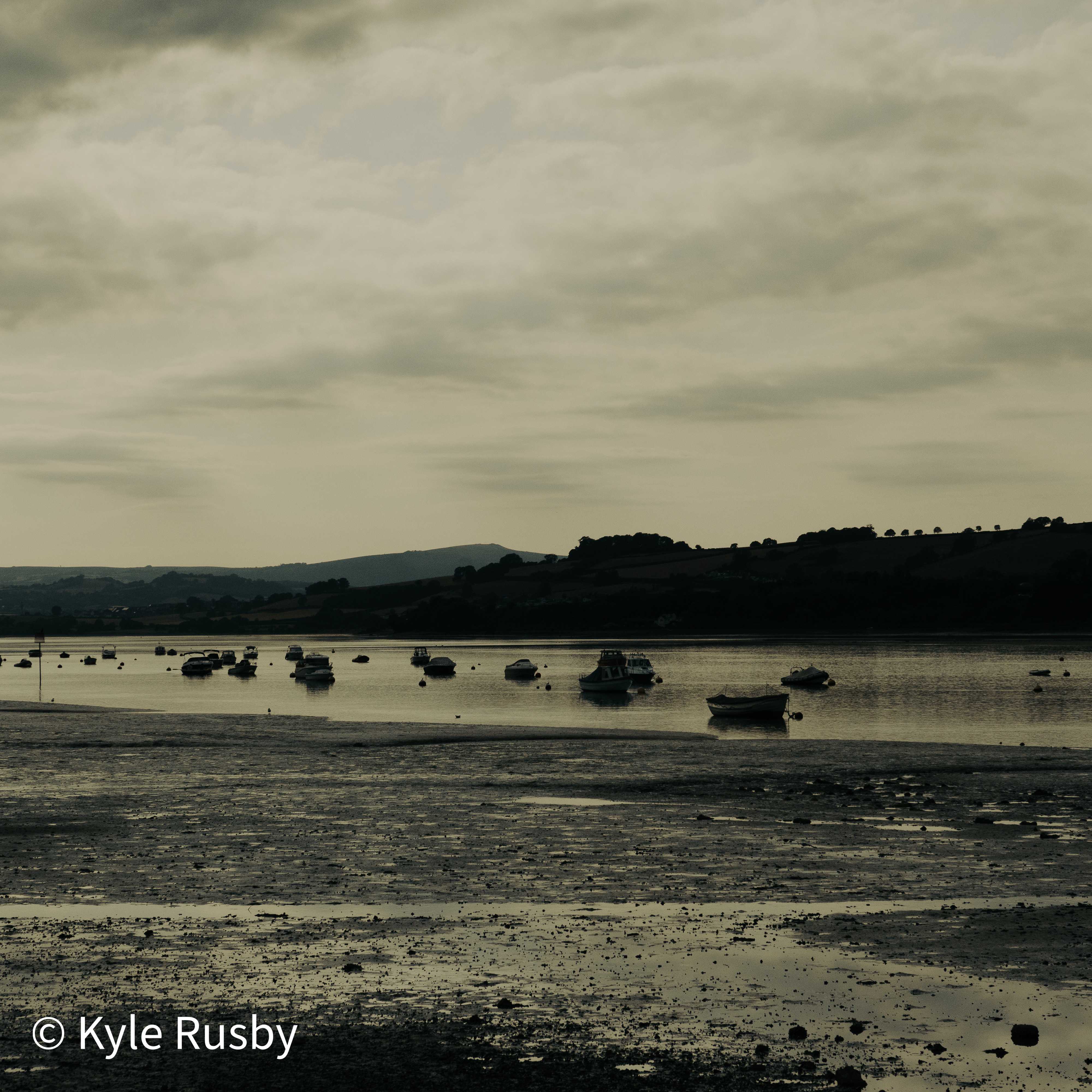 Moored boats sit on their reflections in a thin band of remaining water, the hills beyond gone to silhouette under a broad grey evening sky