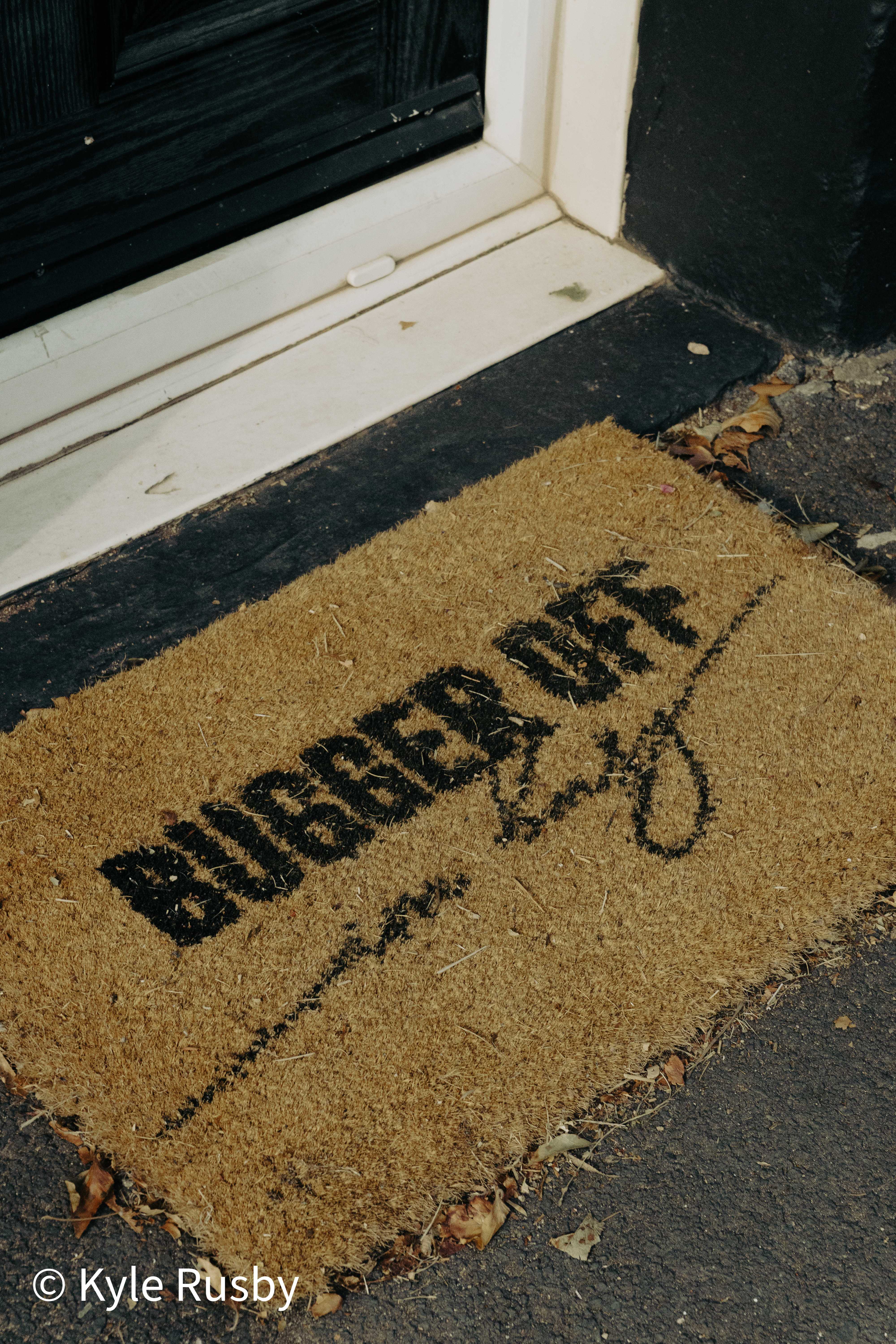 A coir doormat printed with the words BUGGER OFF lies on tarmac at the foot of a black front door