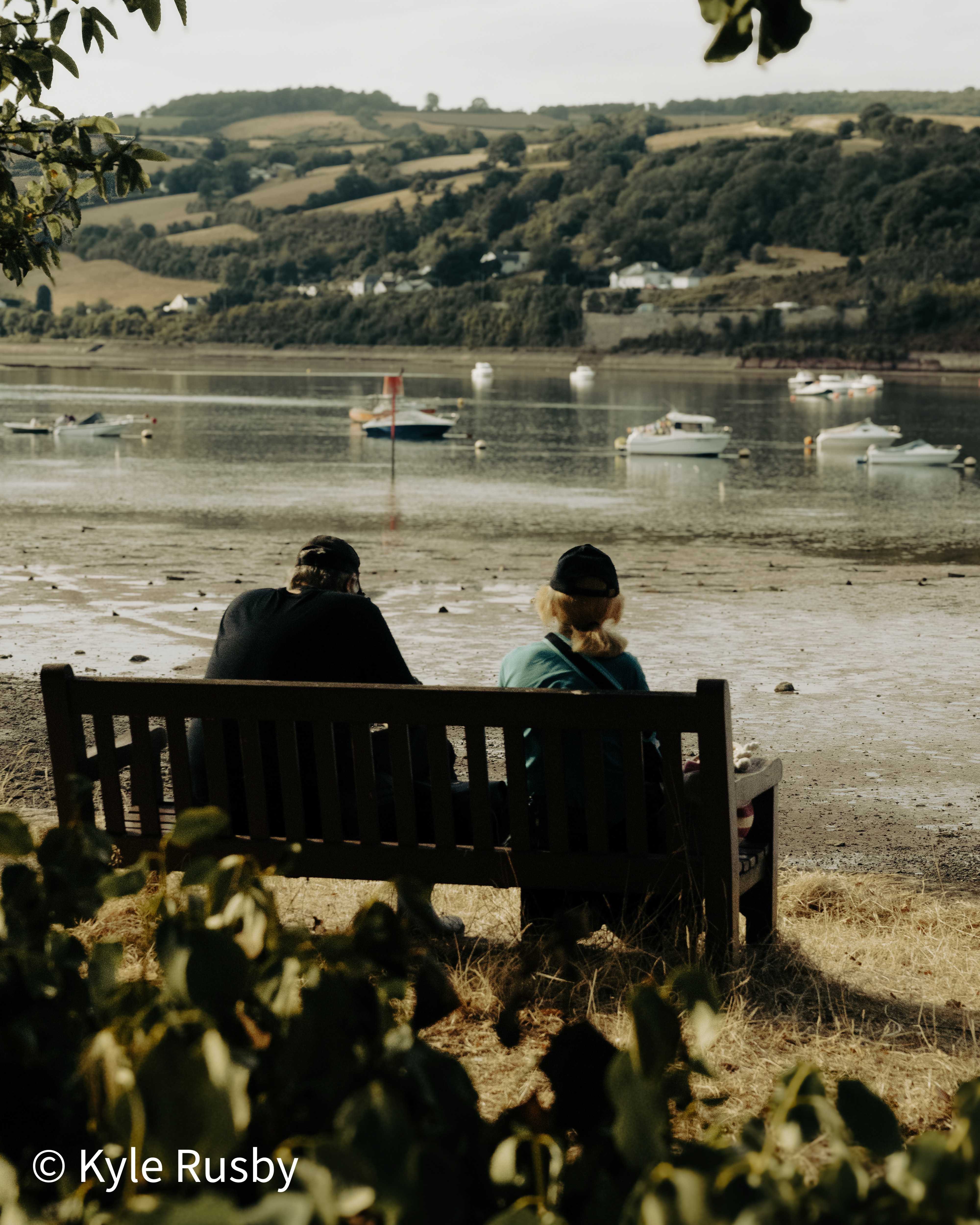 Two people sit on a wooden bench with their backs to the camera, looking out over moored boats and the wooded far bank of an estuary, framed by leaves