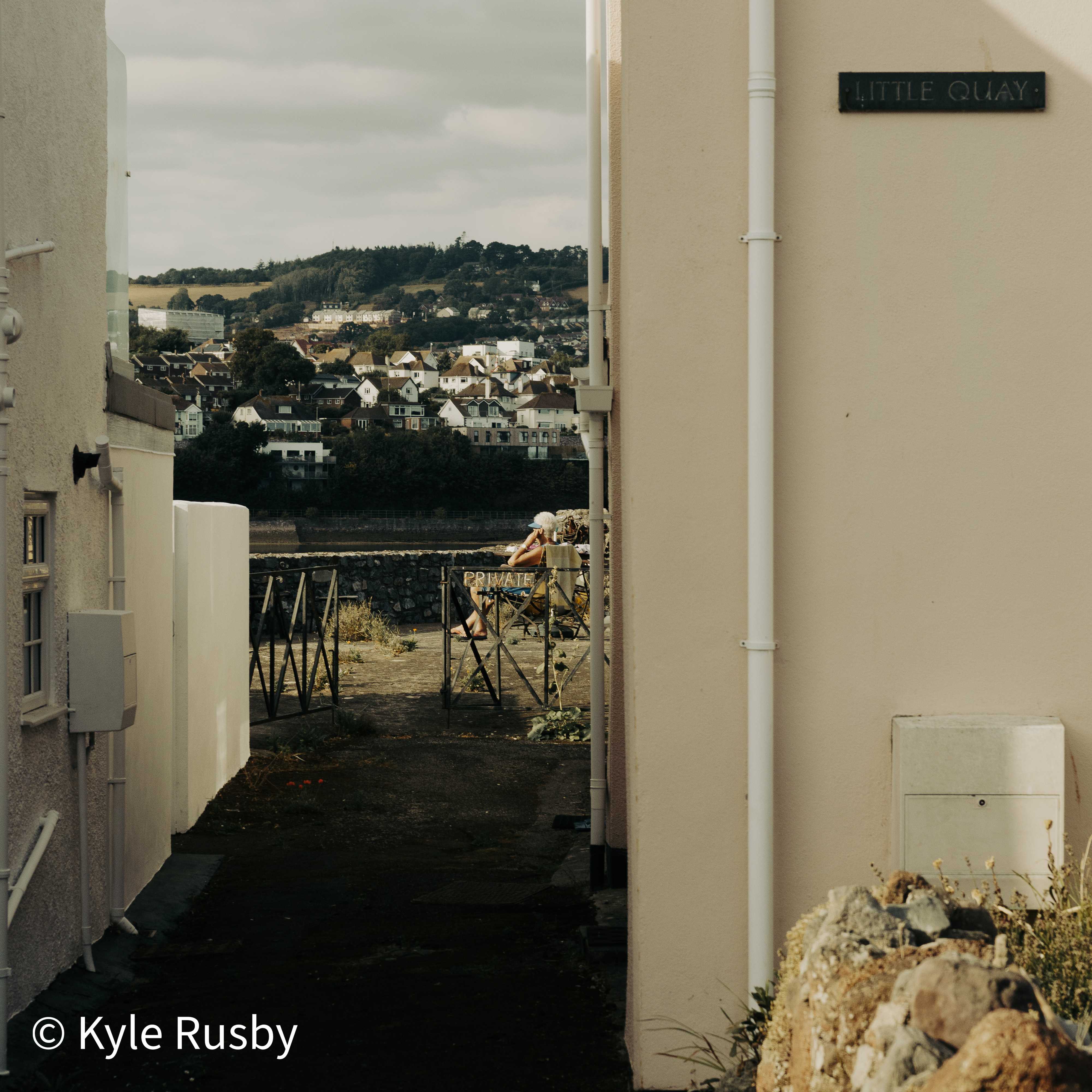 A nameplate reading LITTLE QUAY on a cream wall beside a narrow passage, at the end of which someone sits out in a deckchair behind a PRIVATE sign, the town on its hill across the water