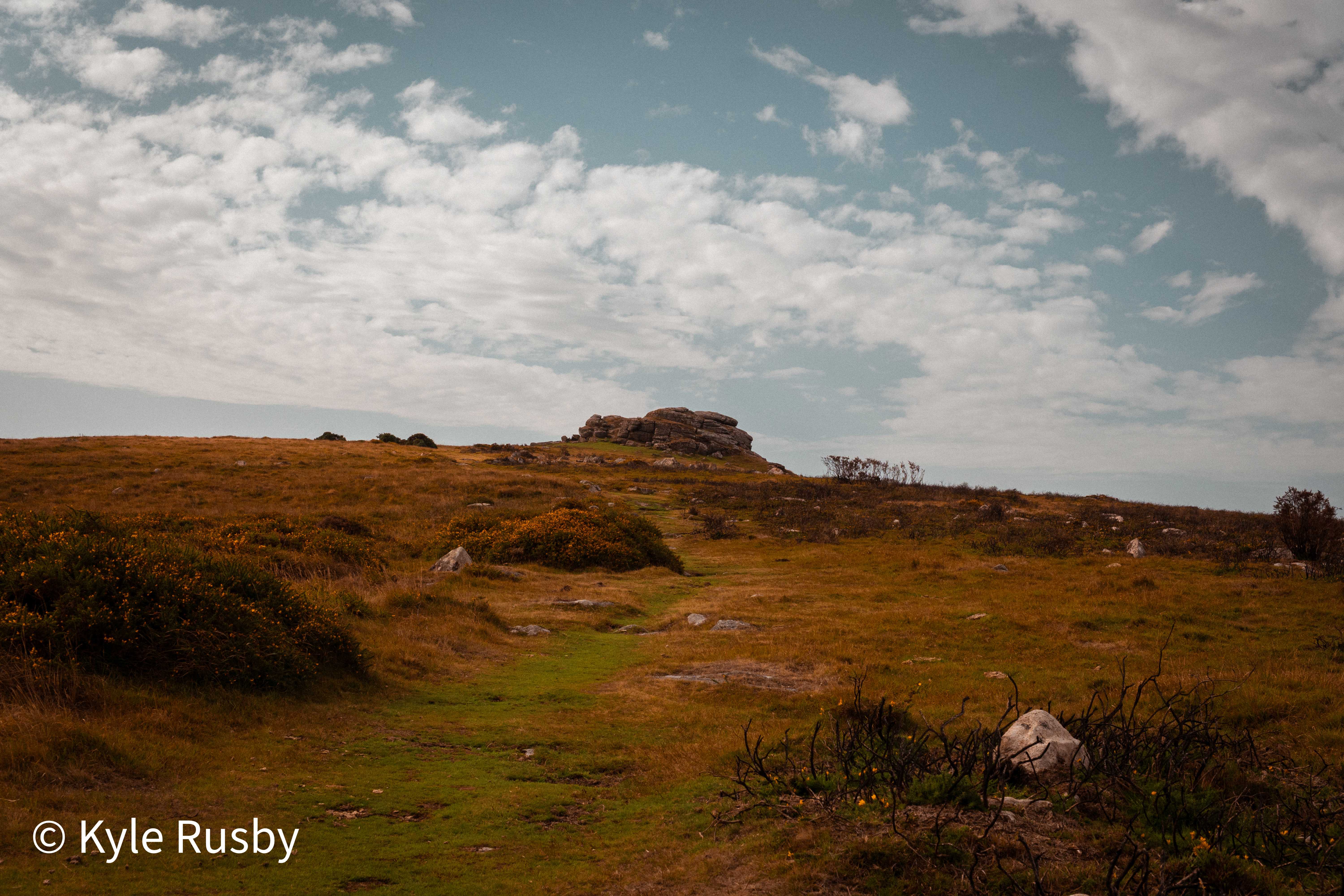 A granite tor sits on the skyline above open moorland of gorse and rust-coloured grass, a green path running up towards it under a wide sky of broken cloud