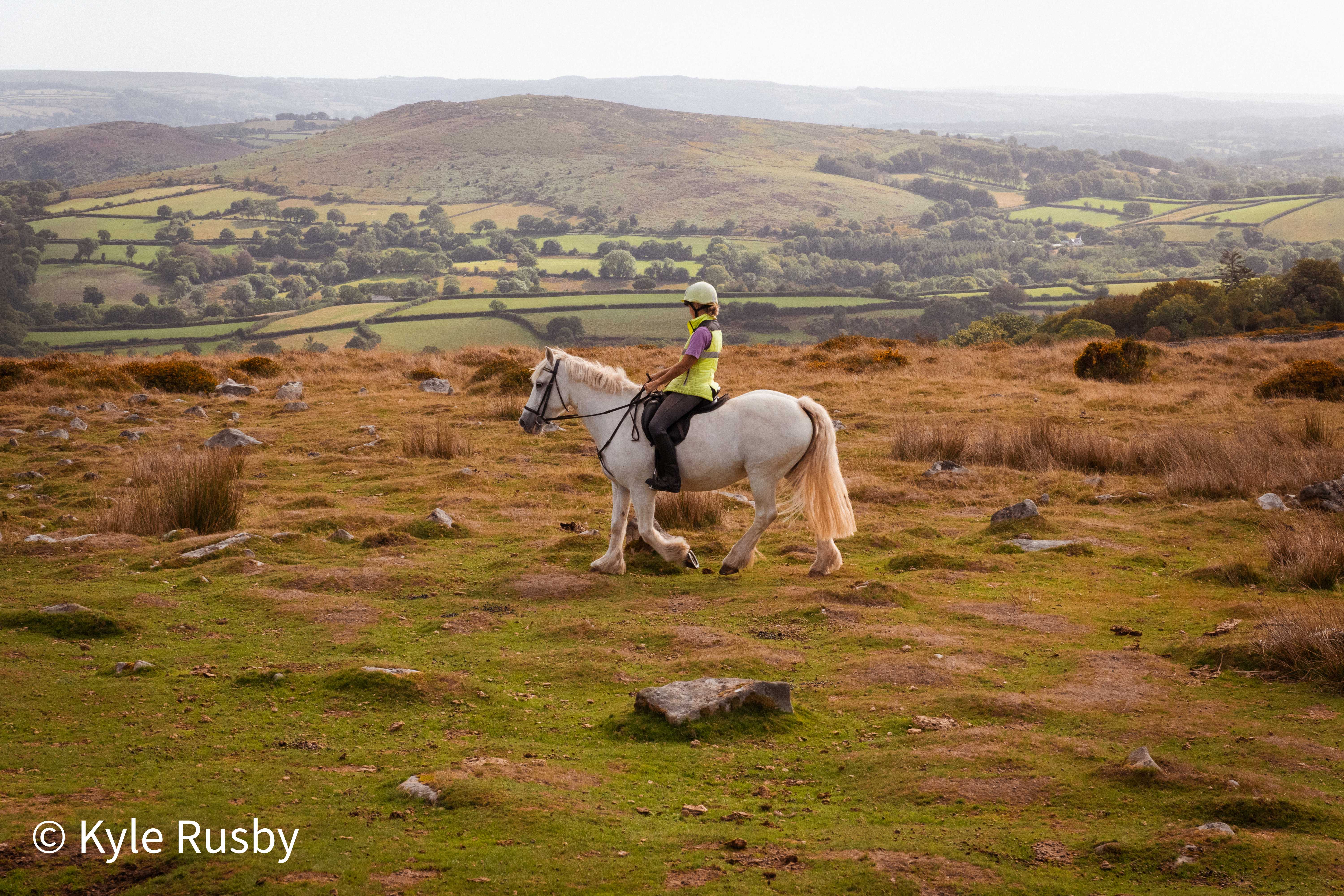 A rider in a hi-vis tabard and white helmet walks a grey cob across open moorland, hills and hedged fields receding into haze behind